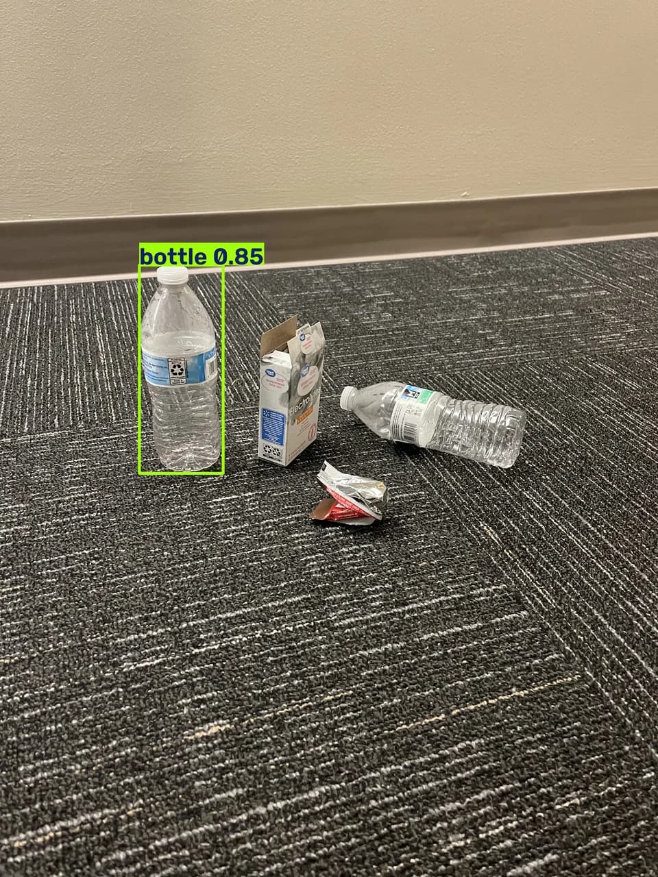 A bottle on carpet among packaging and another bottle, with a green detection box labeled bottle 0.85.